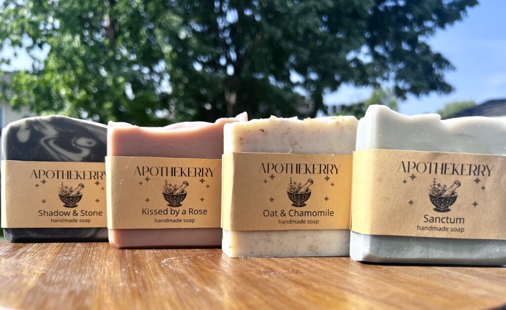 Four ApotheKerry handmade soaps displayed outdoors on a wooden surface, including Shadow & Stone, Kissed by a Rose, Oat & Chamomile, and Sanctum.