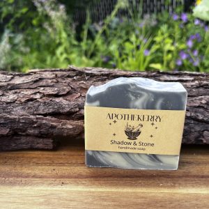 ApotheKerry Shadow & Stone handmade soap with swirling charcoal and clay tones, displayed on a wooden surface with a natural garden background.