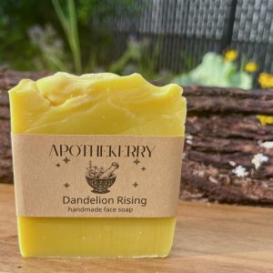 A bright yellow bar of ApotheKerry Dandelion Rising handmade face soap displayed outdoors on a wooden surface with tree bark and soft greenery in the background.