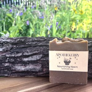 A bar of ApotheKerry Nourishing Neem handmade soap displayed outdoors on a wooden surface beside textured tree bark with a soft garden background.