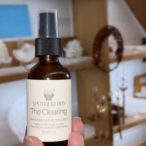 A hand holding an ApotheKerry “The Clearing” herbal mist spray bottle in a softly lit wellness space with shelves and decor blurred in the background.