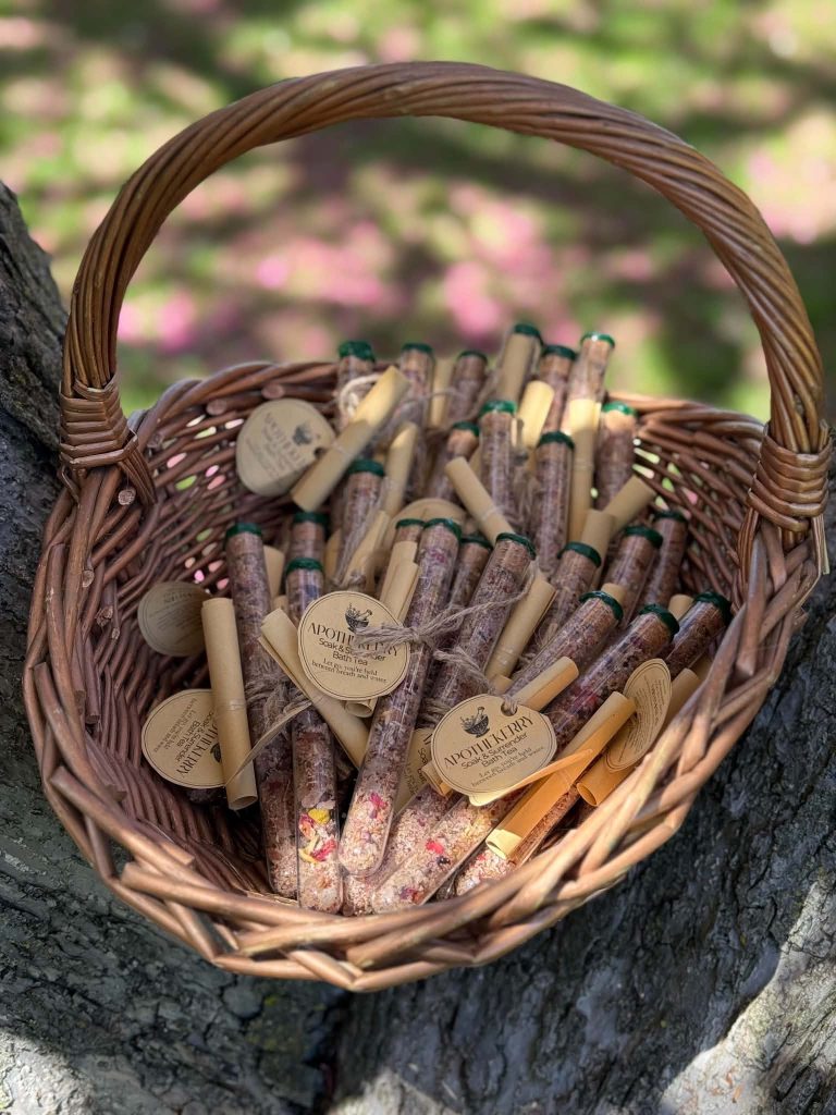 Wicker basket filled with ApotheKerry bath tea tubes resting in a tree outdoors, surrounded by soft pink and green spring foliage.